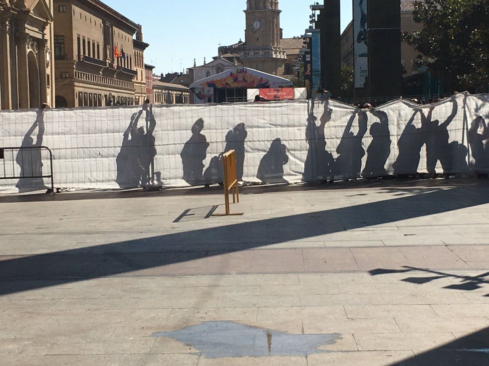 Sombras de los ciudadanos que buscan sortear la malla que rodea la plaza del Pilar para conseguir una imagen de la Virgen del Pilar. Sombras de los ciudadanos que buscan sortear la malla que rodea la plaza del Pilar para conseguir una imagen de la Virgen del Pilar.