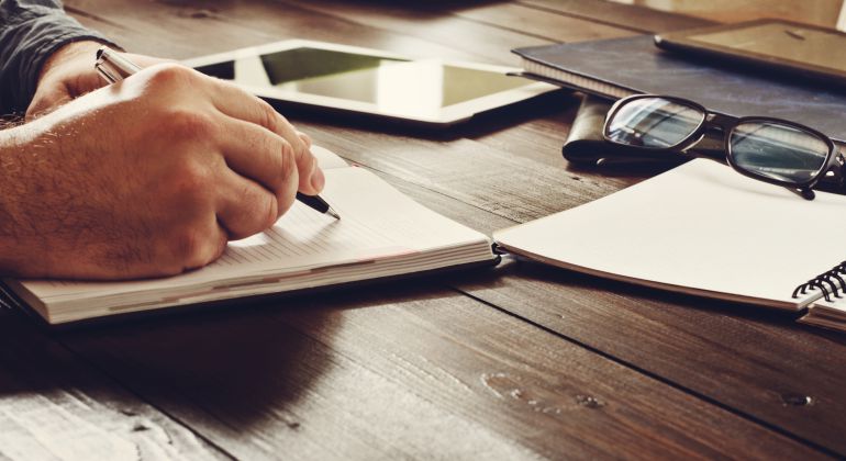 Imagen de un hombre escribiendo junto a un dispositivo digital, varias libretas y un par de gafas. Imagen de un hombre escribiendo junto a un dispositivo digital, varias libretas y un par de gafas.