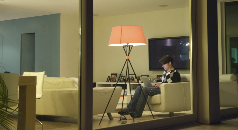 Vista a través de una ventana de un hombre utilizando un ordenador portátil con las luces del salón de su casa encendidas. Vista a través de una ventana de un hombre utilizando un ordenador portátil con las luces del salón de su casa encendidas.