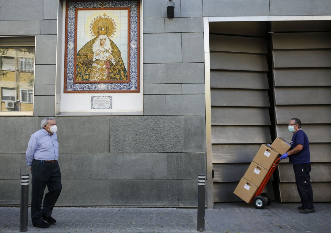 Un hombre con mascarilla pasea por Sevilla, cerca, otro hombre descarga cajas de material también debidamente protegido Un hombre con mascarilla pasea por Sevilla, cerca, otro hombre descarga cajas de material también debidamente protegido