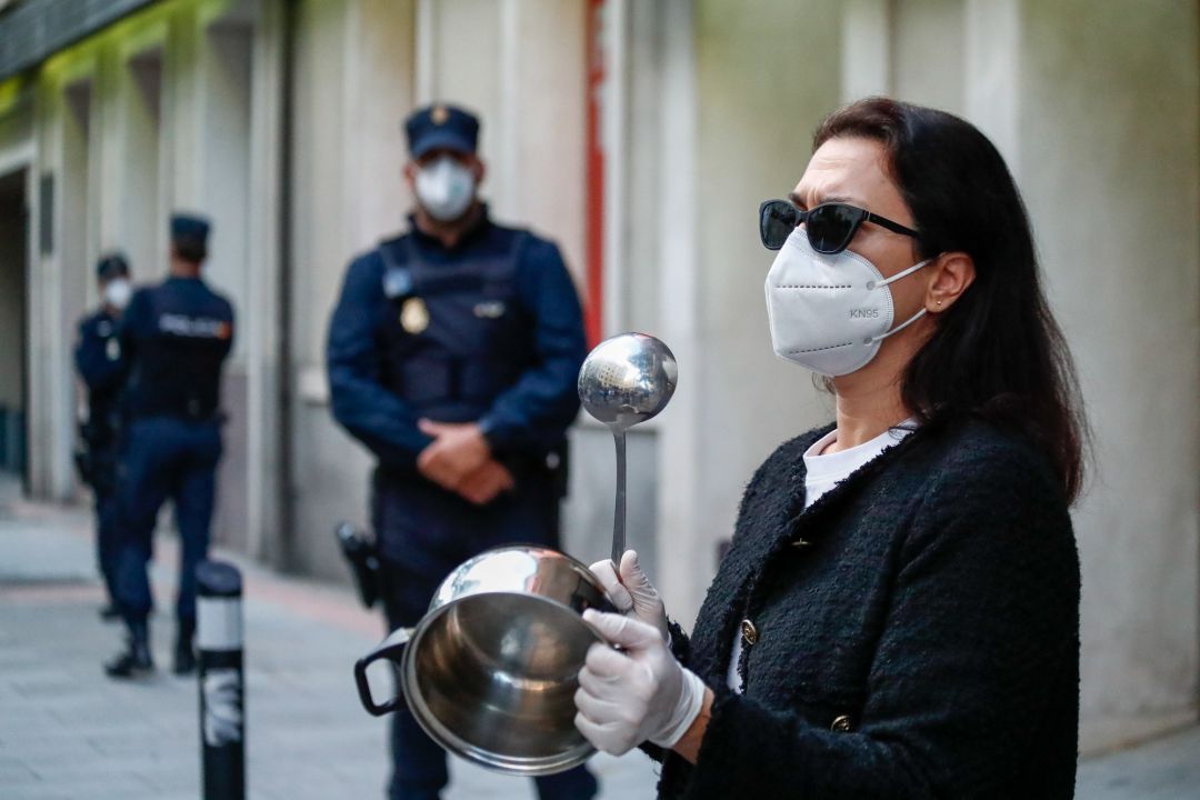 Una mujer con mascarilla golpea una cacerola delante de varios Policías Nacionales. Una mujer con mascarilla golpea una cacerola delante de varios Policías Nacionales.