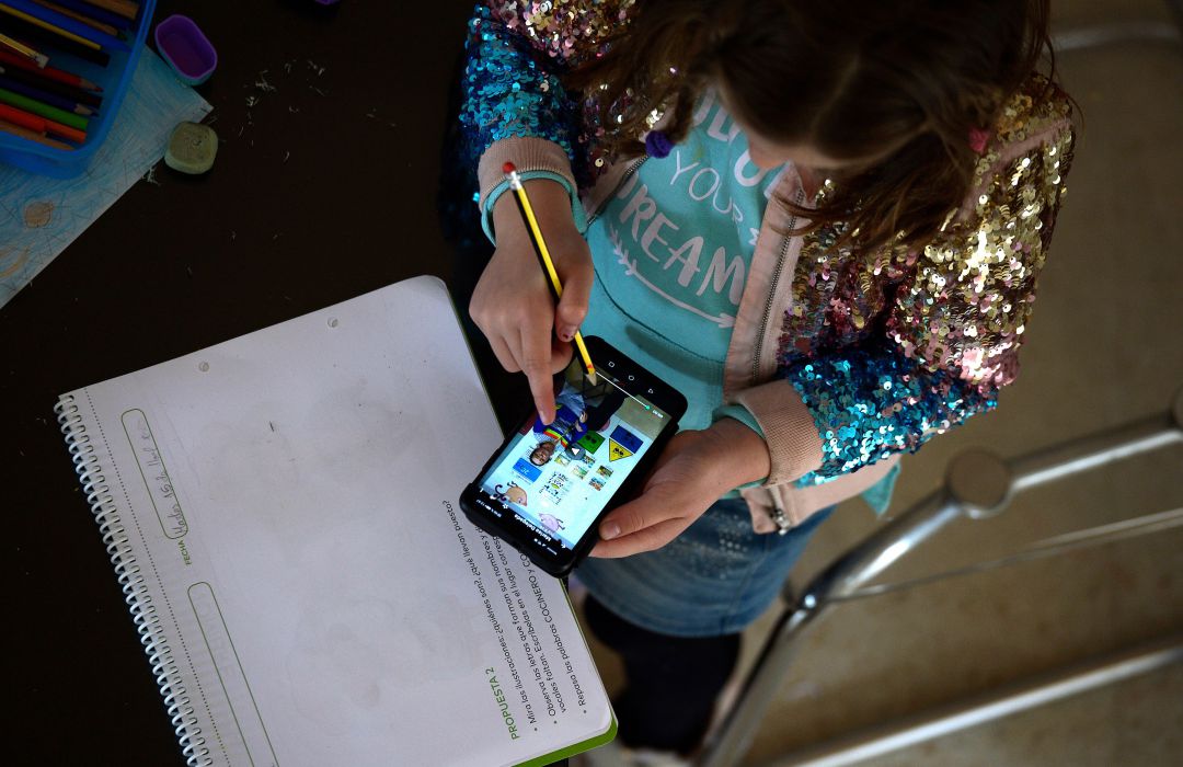 Una menor estudiante de primaria hace deberes mientras sigue las explicaciones de su profesora en vídeo a través de teléfono móvil. Una menor estudiante de primaria hace deberes mientras sigue las explicaciones de su profesora en vídeo a través de teléfono móvil.