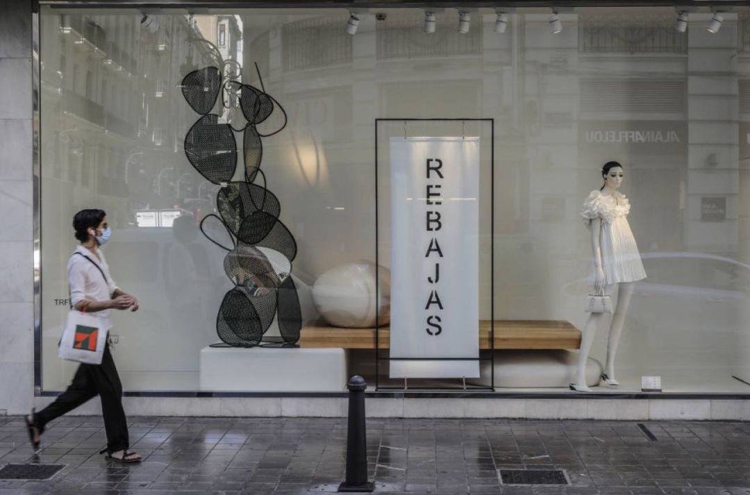 Una mujer protegida con mascarilla pasa al lado del escaparate de una tienda donde se observan carteles indicativos de rebajas en València Una mujer protegida con mascarilla pasa al lado del escaparate de una tienda donde se observan carteles indicativos de rebajas en València
