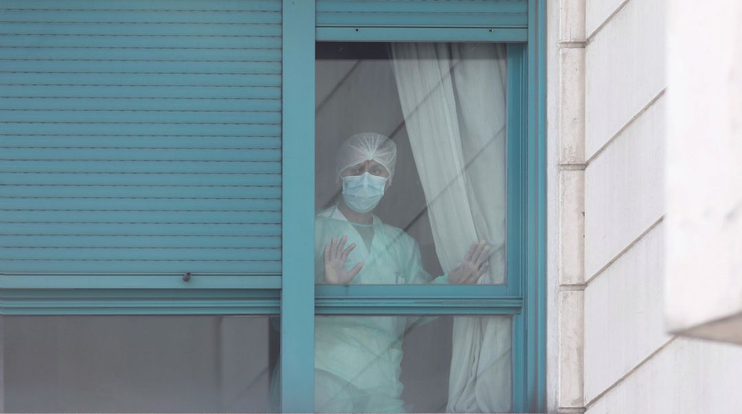 Una trabajadora del personal sanitario protegida con mascarilla se asoma por una de las ventanas de una residencia. Una trabajadora del personal sanitario protegida con mascarilla se asoma por una de las ventanas de una residencia.