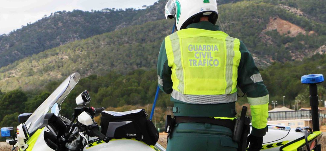Un agente de la Guardia Civil de Tráfico, de espaldas, en una carretera junto a su moto y una montaña al fondo. Un agente de la Guardia Civil de Tráfico, de espaldas, en una carretera junto a su moto y una montaña al fondo.