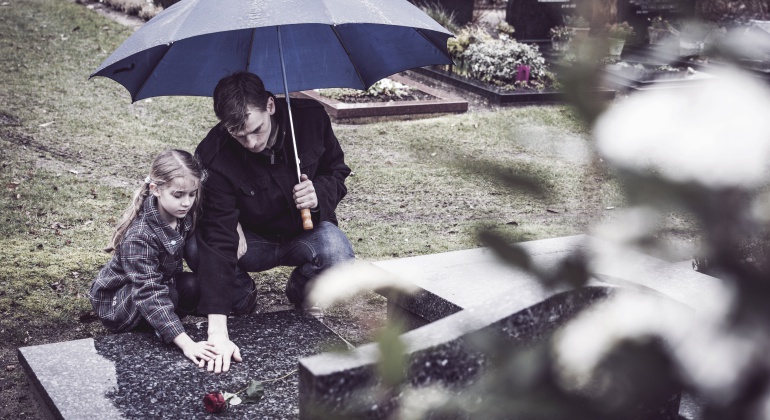 Un padre y su hija depositan una flor en una tumba de un cementerio.