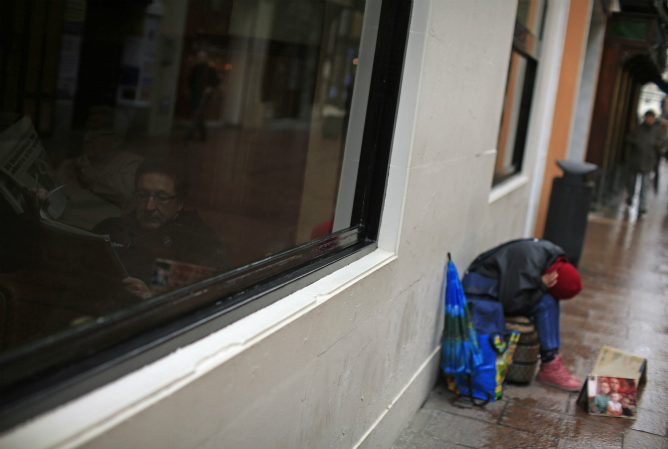 Un hombre lee un periódico en el interior de un bar mientras un mendigo está pidiendo en una calle de Sevilla