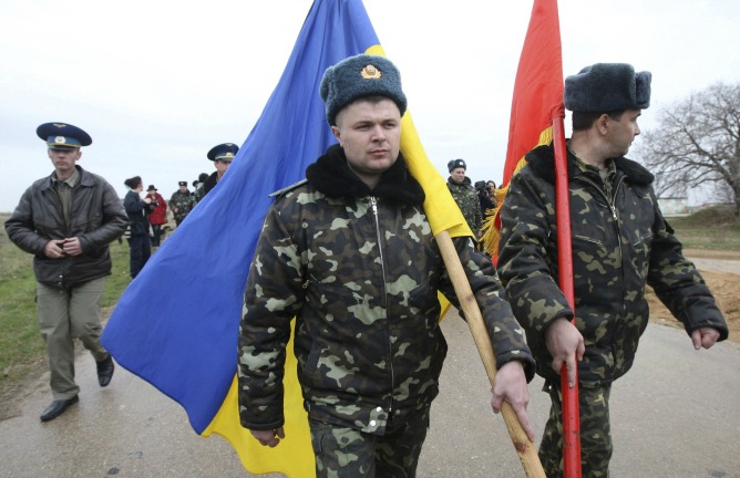 Tropas ucranianas, con banderas de Ucrania, marchan mientras hombres armados y uniformados bloquean la entrada de la base aérea de Bilbek