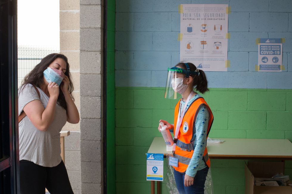 Una trabajadora de Protección Civil a las puertas de un colegio electoral en Galicia espera a que una votante se coloque su segunda mascarilla para ofrecerle gel hidroalcohólico antes de entrar. Una trabajadora de Protección Civil a las puertas de un colegio electoral en Galicia espera a que una votante se coloque su segunda mascarilla para ofrecerle gel hidroalcohólico antes de entrar.